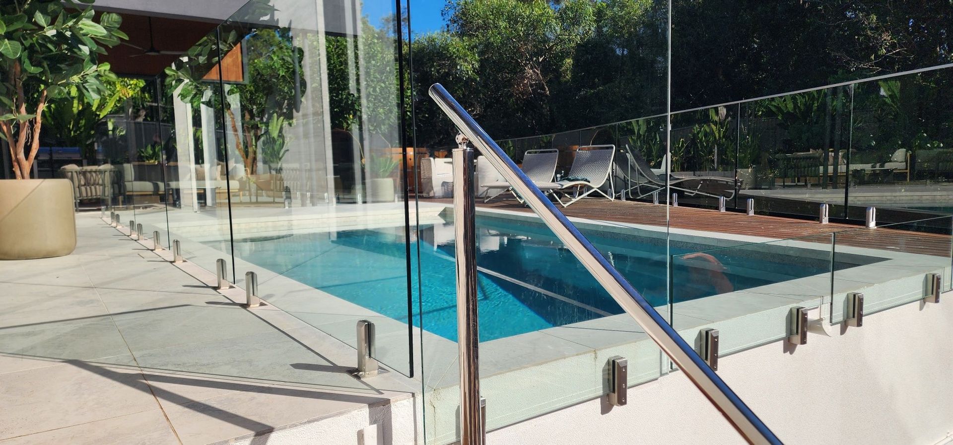 Outdoor swimming pool with glass safety fence, stainless handrail, and surrounding patio under bright sunlight