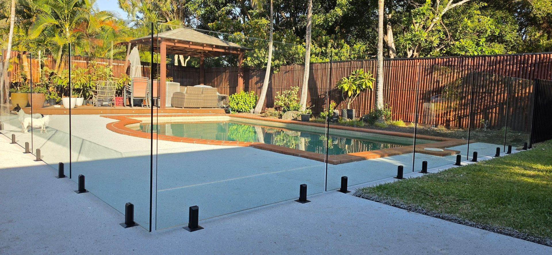 Empty backyard with a rectangular pool, gravel patio, black posts, and a covered seating area surrounded by trees.