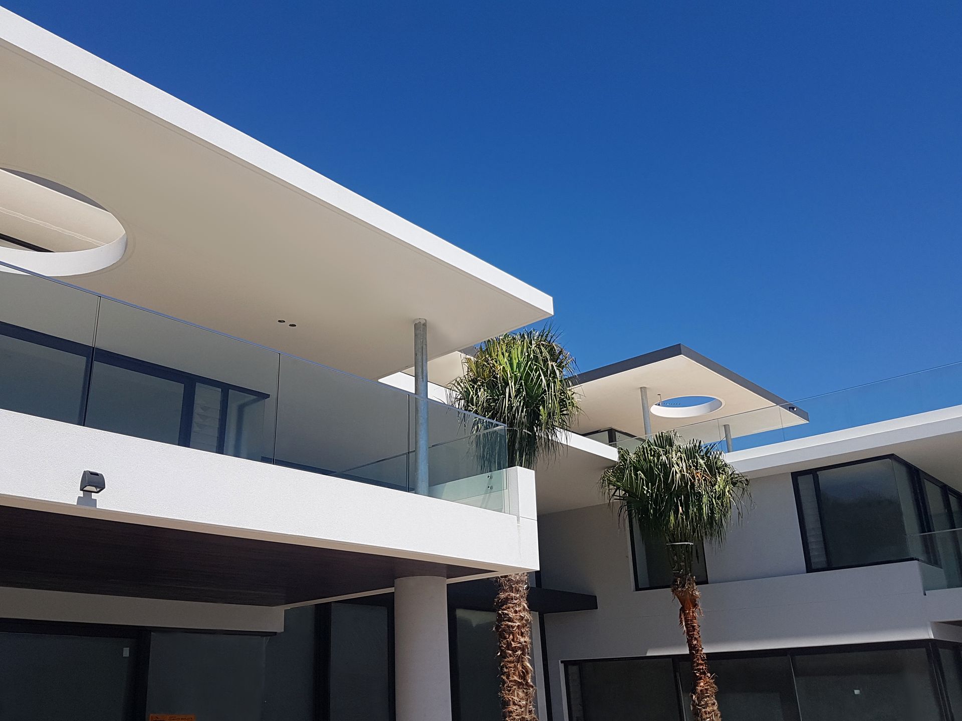 Modern white buildings with glass balconies and palm trees against a blue sky.