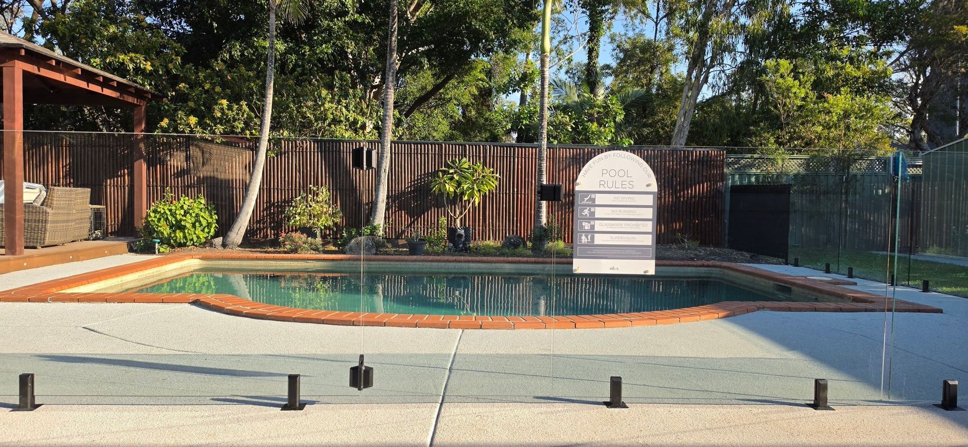 Backyard swimming pool with patio, wooden fence, and two white lounge chairs under trees