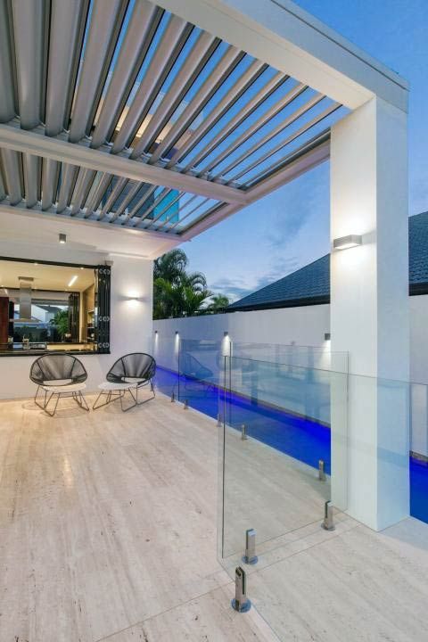 Patio with gray pergola, pool with blue light, glass railing. Two chairs and a window.