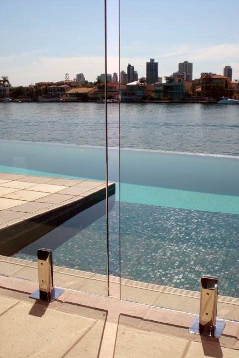 Infinity pool with glass barrier overlooking a river and city skyline on a sunny day.