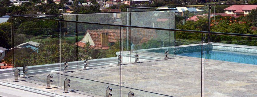 Rooftop pool with glass railing overlooking a town. Blue water, gray concrete, trees, and buildings.
