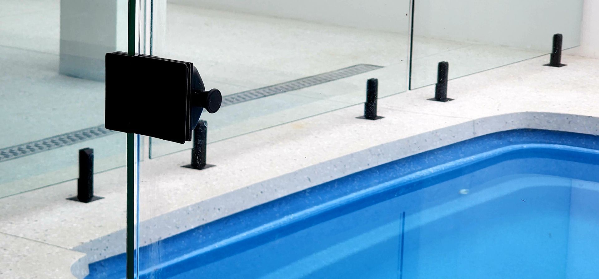 A glass pool fence with black posts and a black gate latch next to a blue pool.