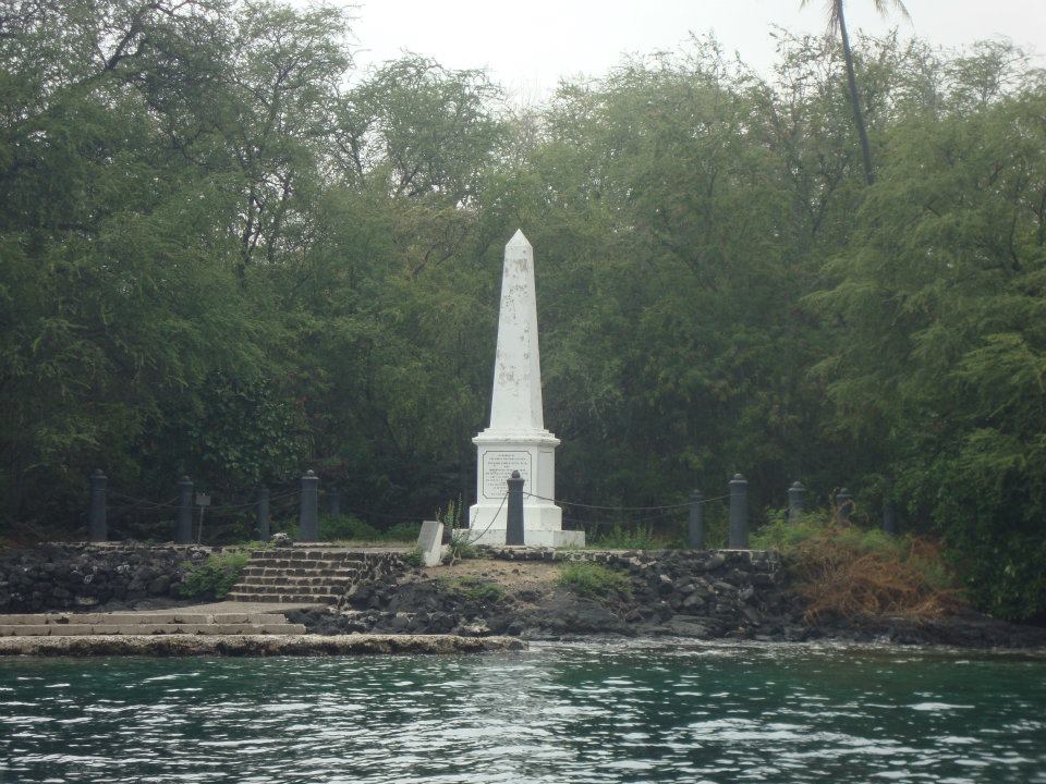 White monument on a rocky shoreline, backed by dense green trees, viewed across calm water.