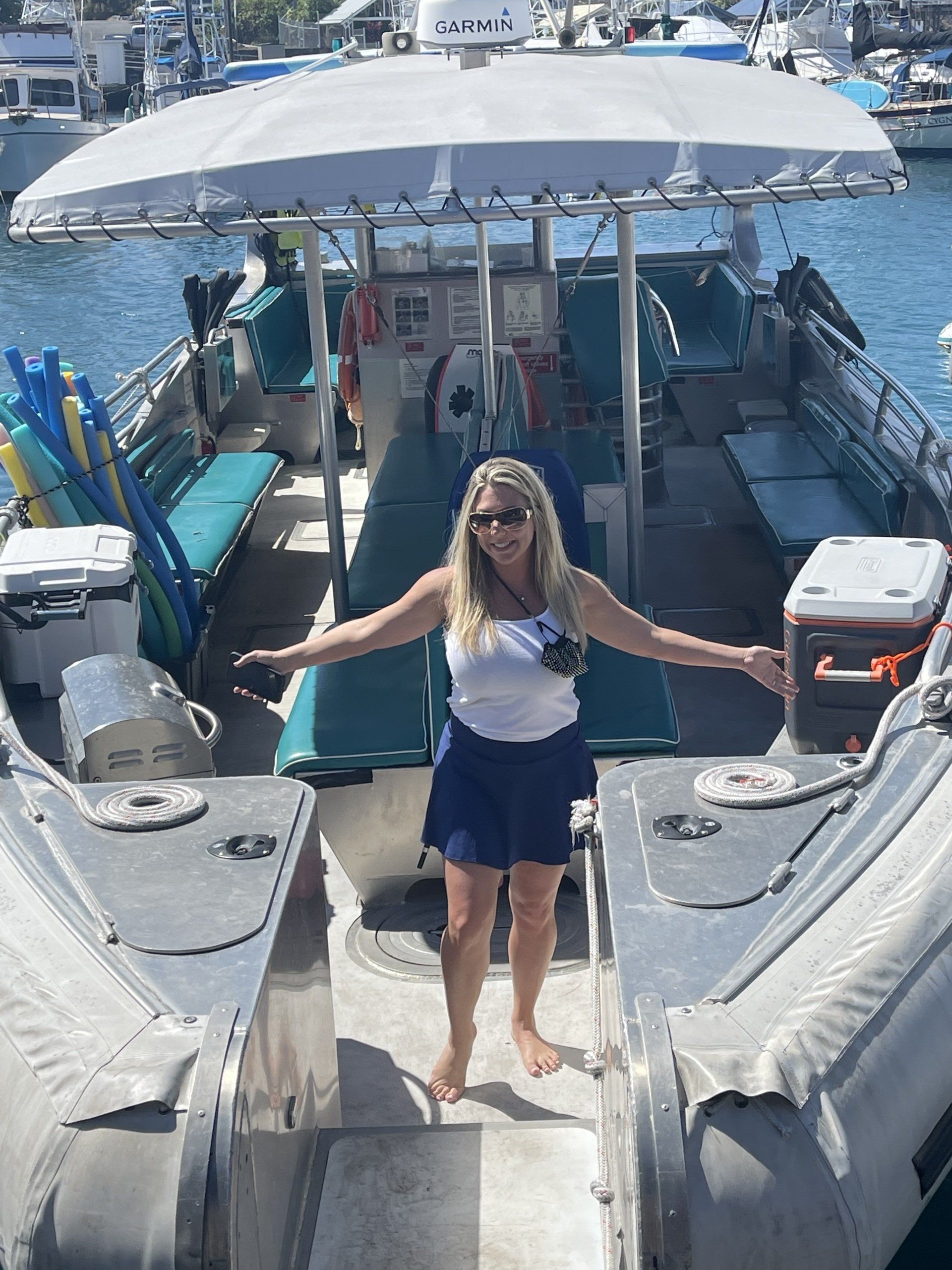 Woman standing on the bow of a boat, arms outstretched, with blue water and docked boats behind her.