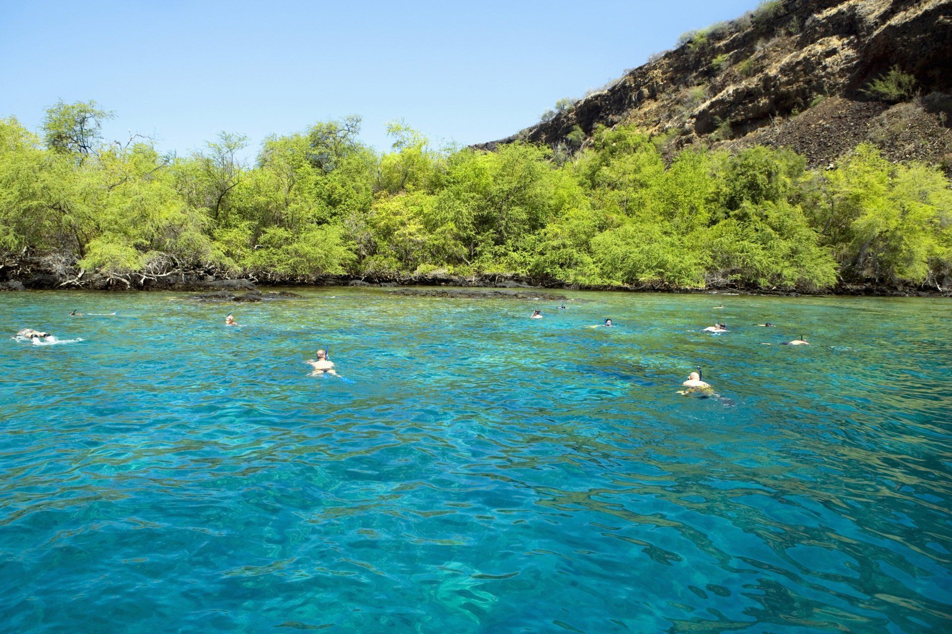 Turquoise tropical water with swimmers near a green shoreline and rocky hill under a clear blue sky