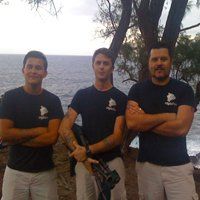 Three men in dark blue shirts stand with arms crossed by the sea, one holding a camera