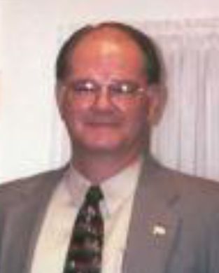 Man in a gray suit and patterned tie, smiling in front of a light curtain backdrop
