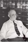 Man in white shirt and tie seated at a desk with shelves of jars behind him
