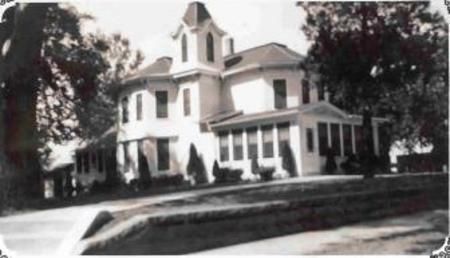 Black-and-white photo of a large Victorian-style house with a porch and trees in front