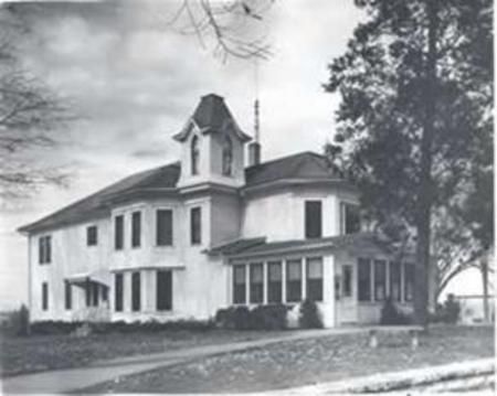 Historic white house with a tower and porch, surrounded by trees on a grassy lawn.
