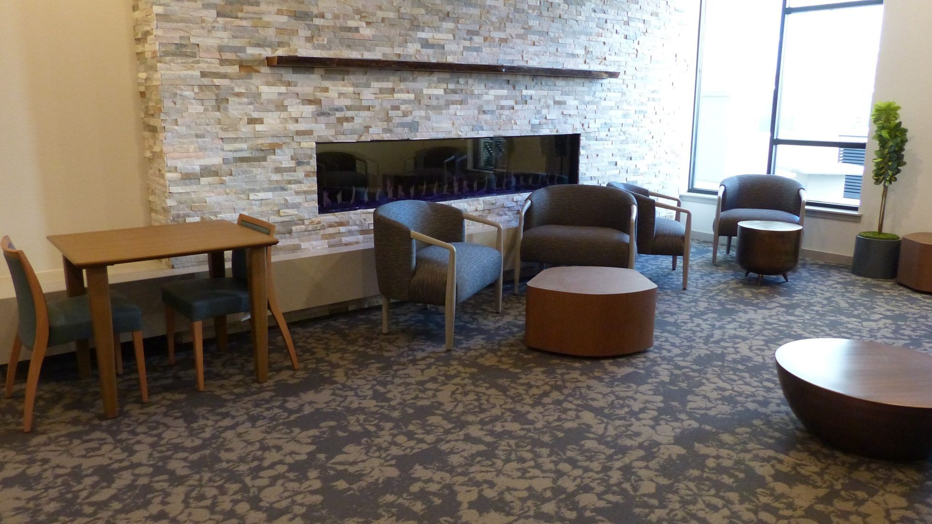 Lounge with stone fireplace, patterned carpet, and scattered chairs and tables by a bright window.