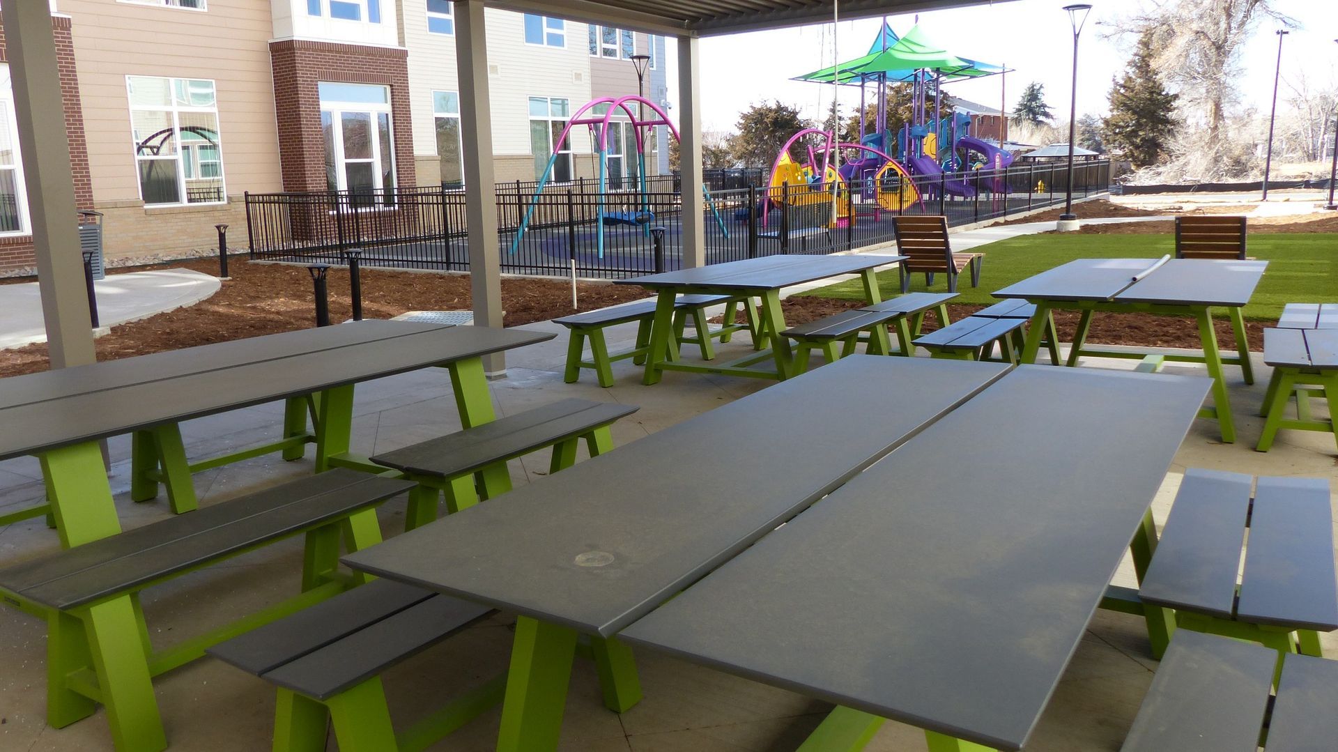Outdoor picnic tables under a covered patio beside a playground with colorful equipment in the background