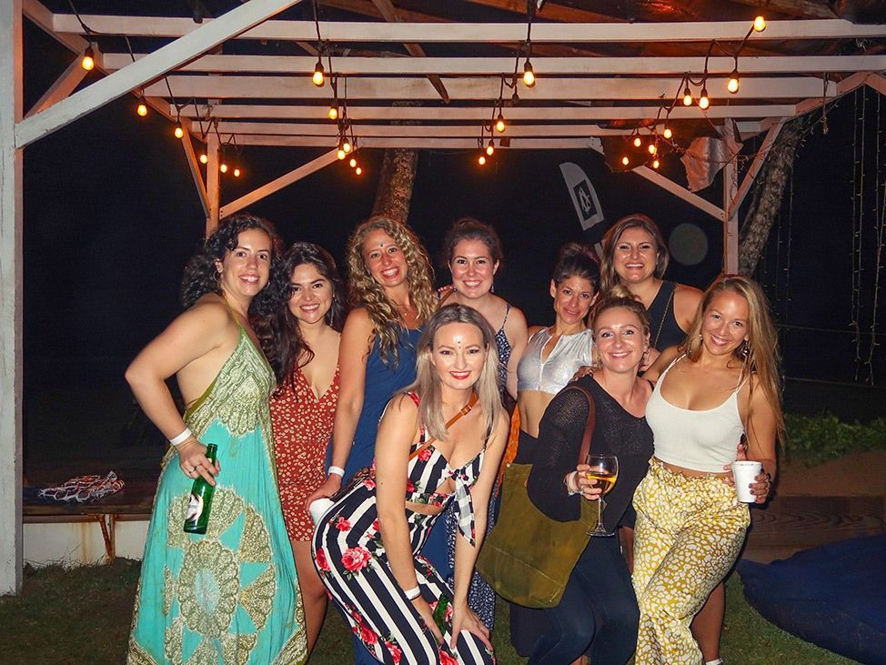 Group of people smiling and posing under a lit canopy at night, holding drinks
