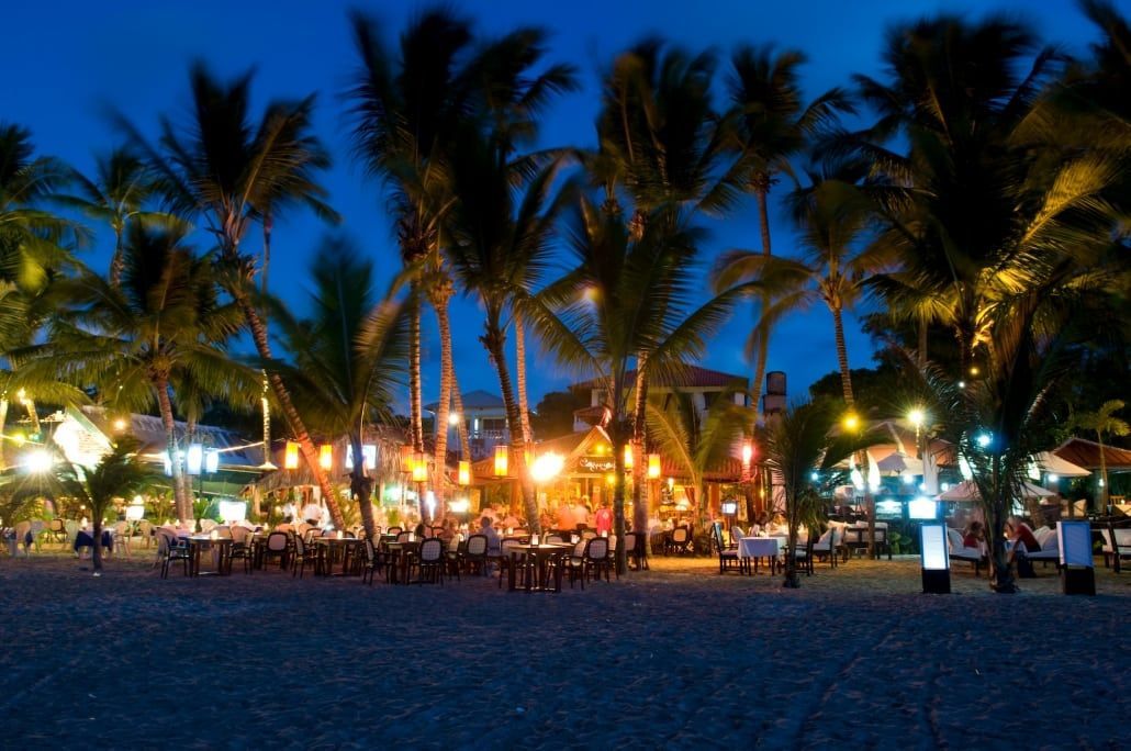 Palm-lined beach at dusk with bright lights, people gathered, and a blue evening sky