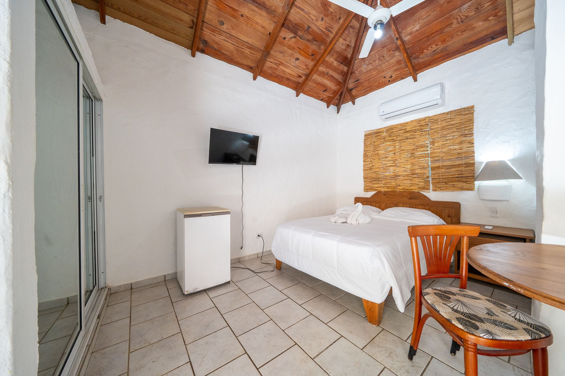 Small rustic bedroom with white bed, wall TV, tiled floor, wood ceiling, and chair by a window blind