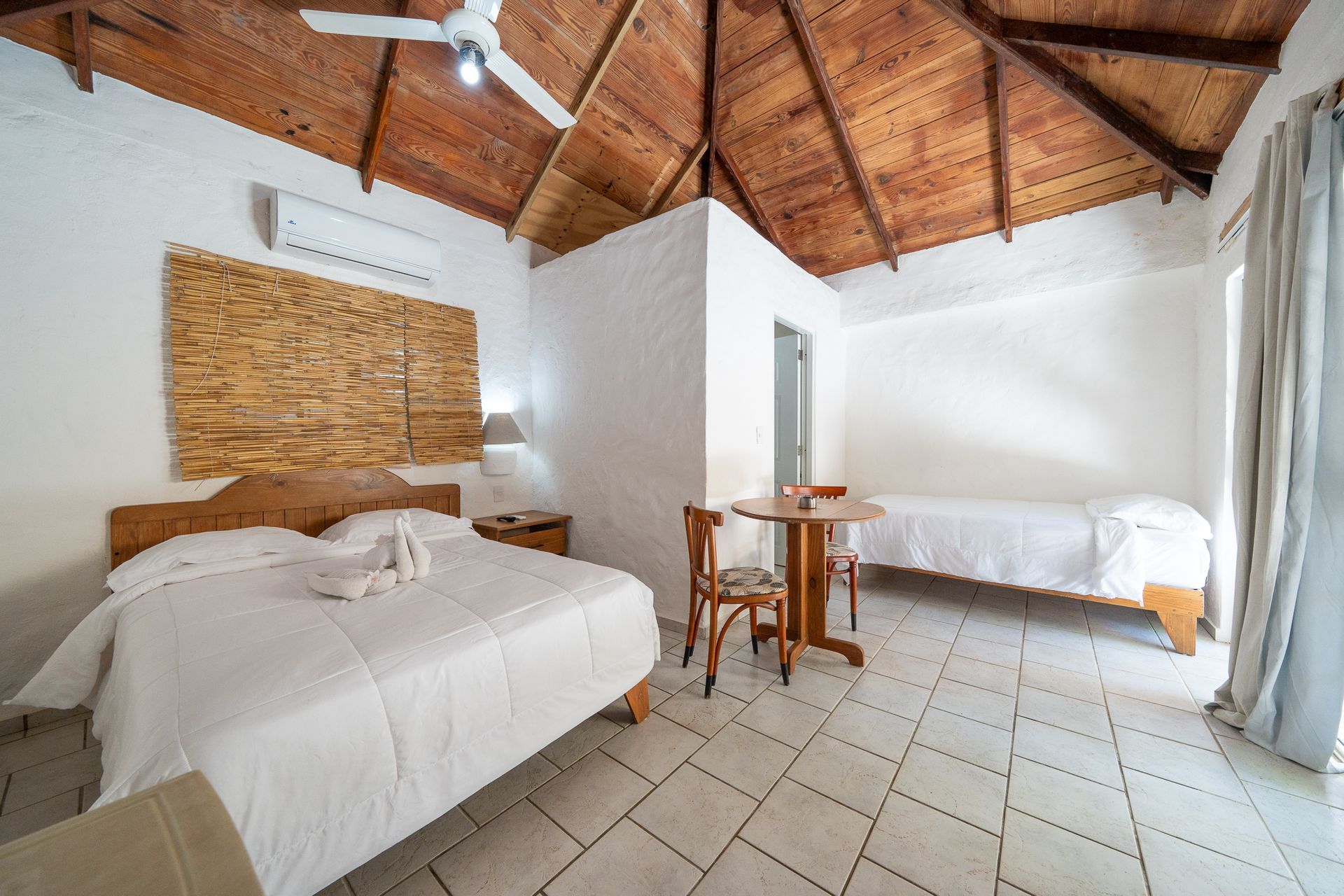 Rustic bedroom with white walls, wooden vaulted ceiling, two beds, and a small table by the window