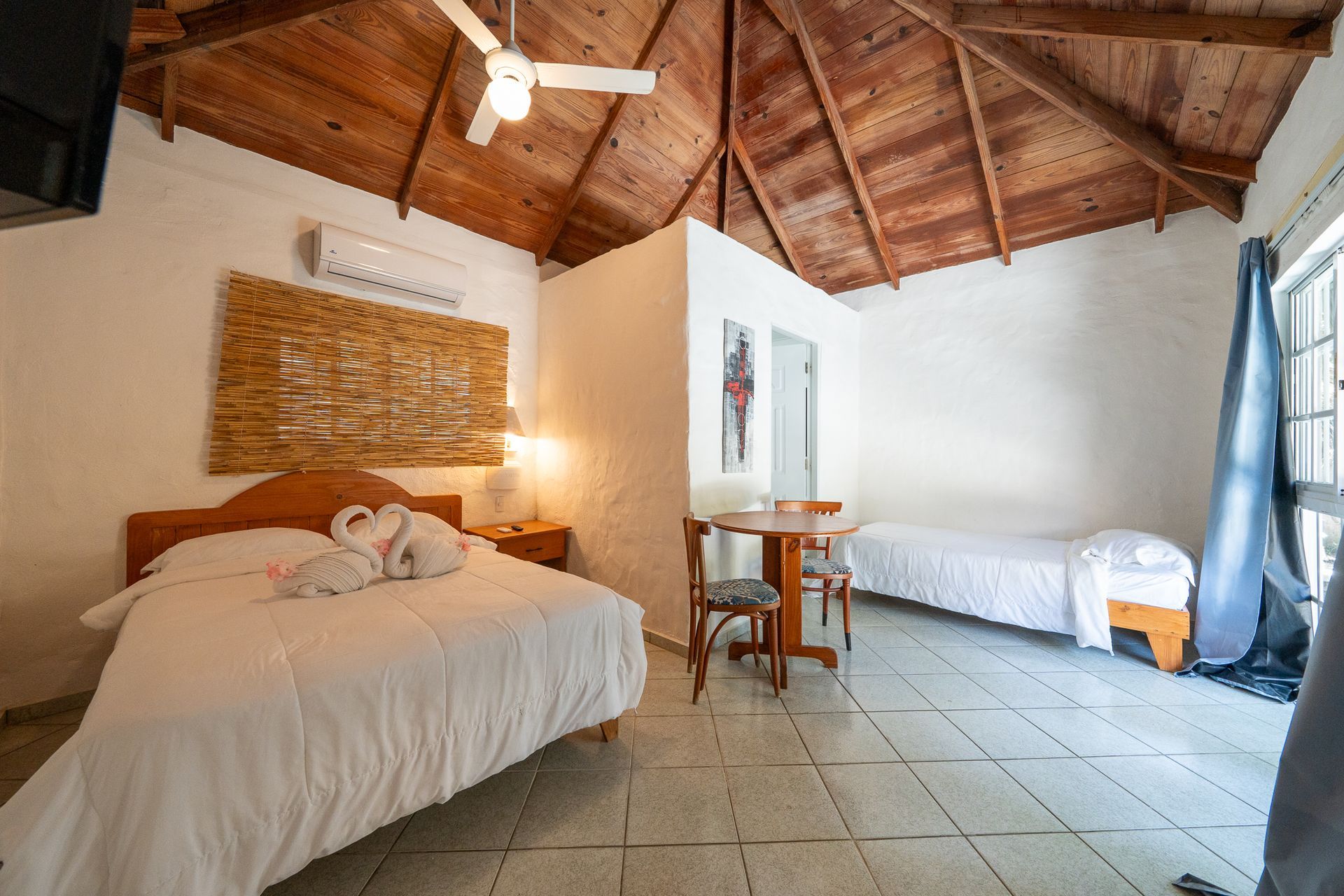Rustic bedroom with two white beds, tiled floor, wood ceiling, and blue curtains