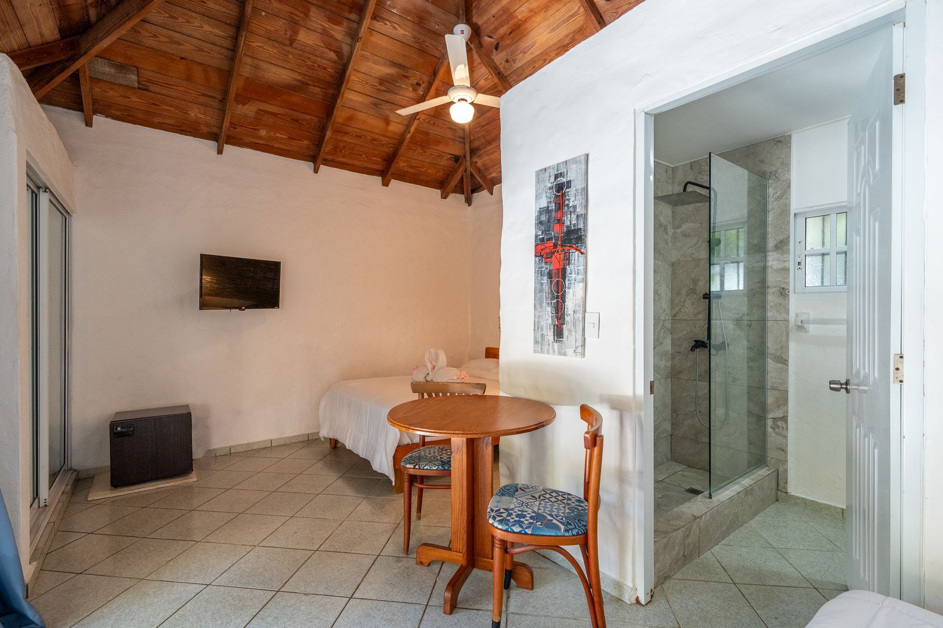 Small room with wooden ceiling, twin beds, table and chairs, and a glass-walled bathroom.