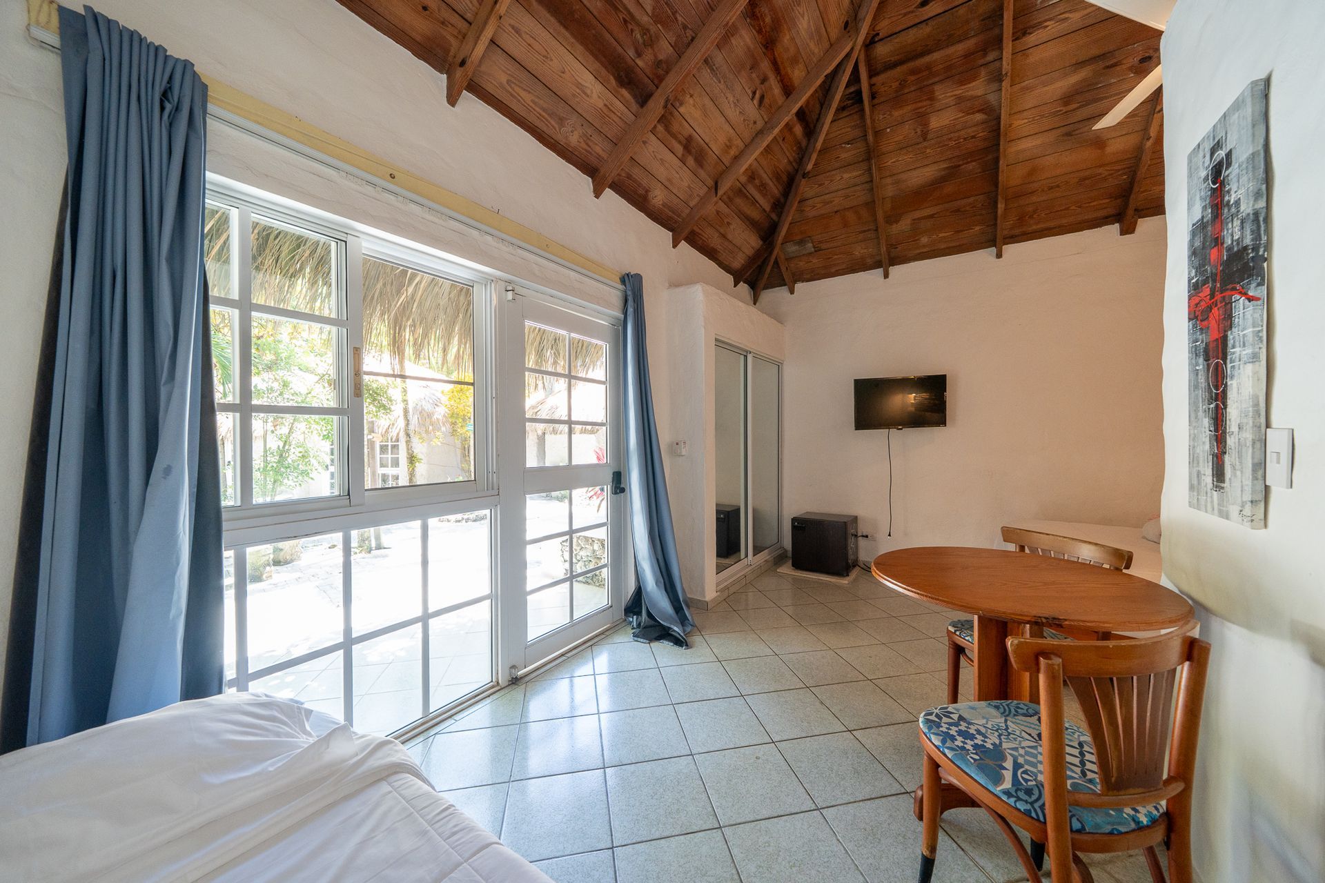 Bright room with large windows, tiled floor, wooden vaulted ceiling, and small dining table with chairs.