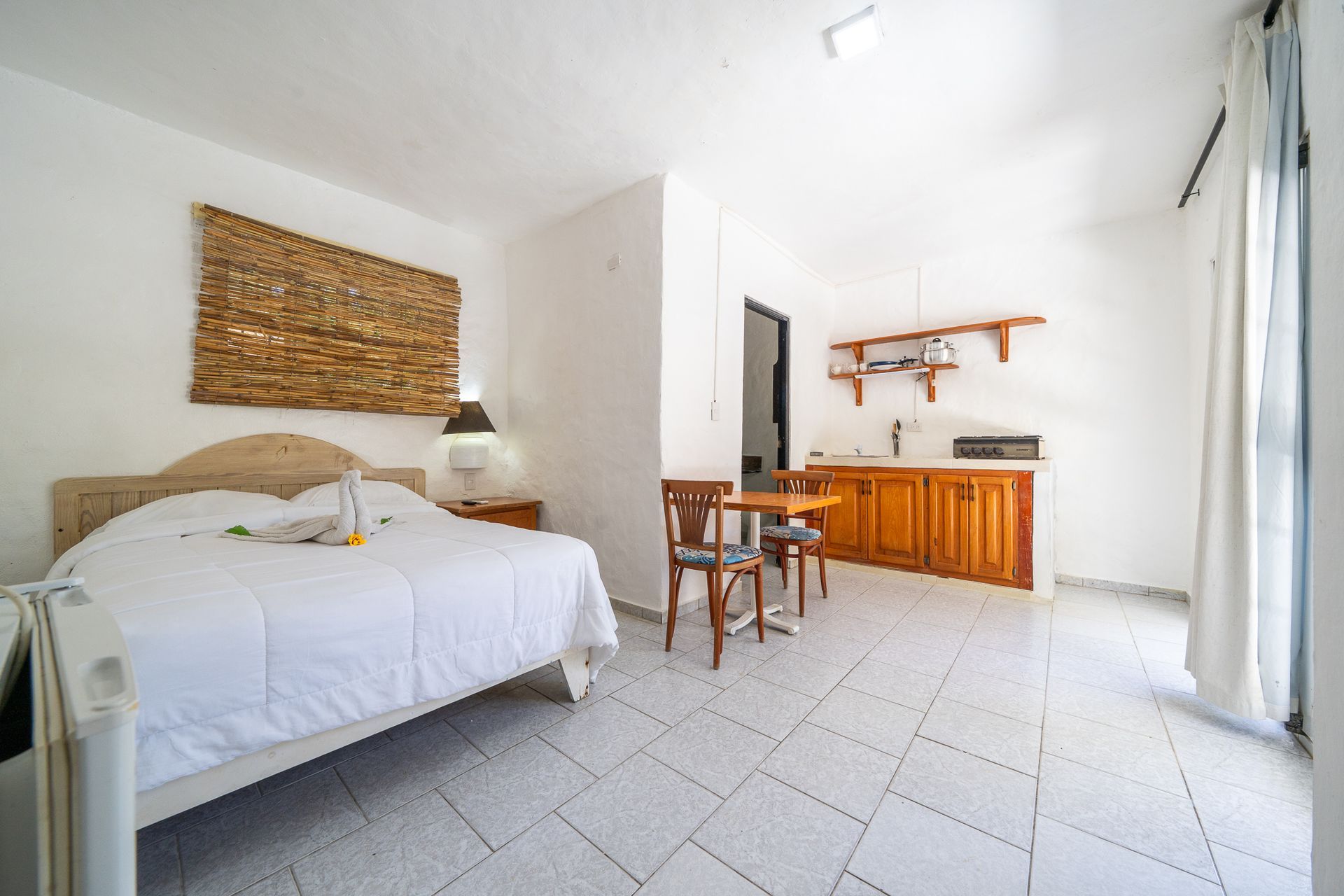 Bright bedroom with white bed, wicker wall art, small desk, and kitchenette by sliding glass doors