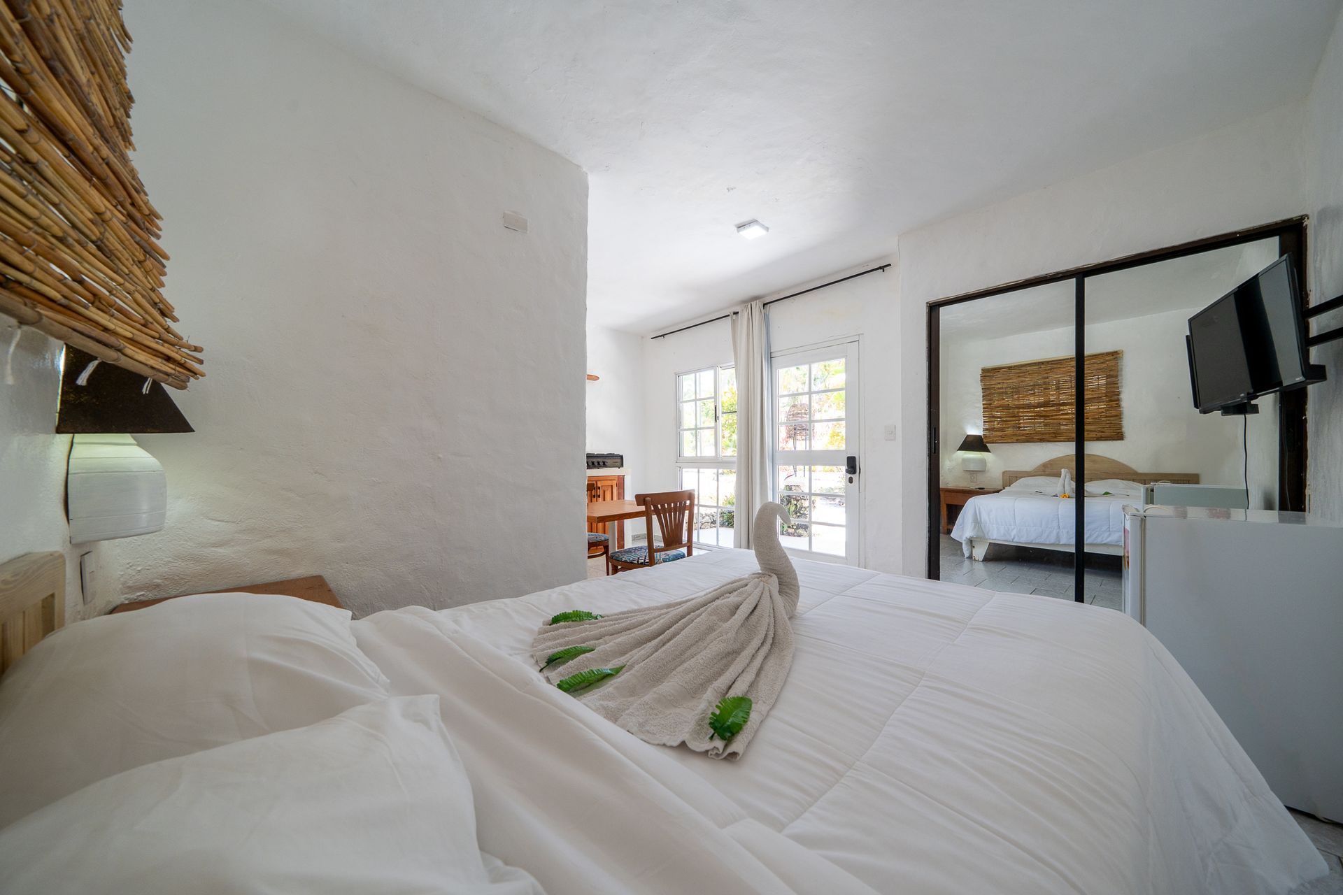 Bright bedroom with white bed, large mirror, desk, and balcony doors letting in natural light.