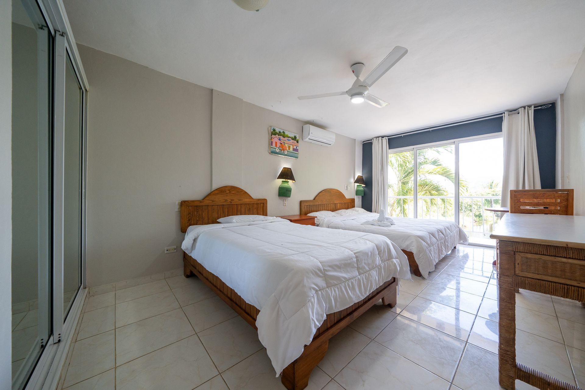 Bright bedroom with two wooden beds, tiled floor, ceiling fan, and sliding doors to a balcony.