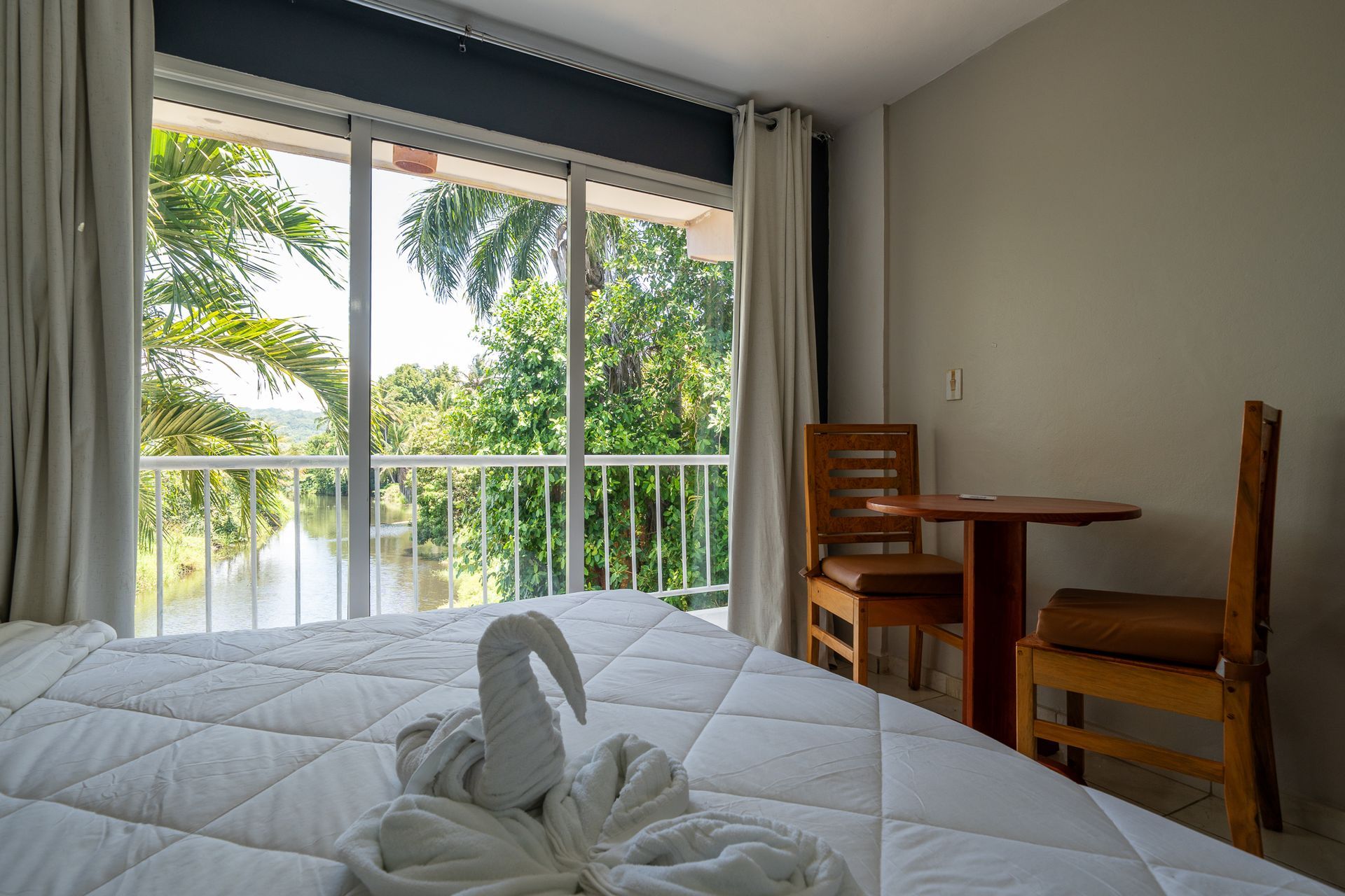 Bright bedroom with a neatly made bed, wooden chairs, and a large window overlooking palm trees and water.