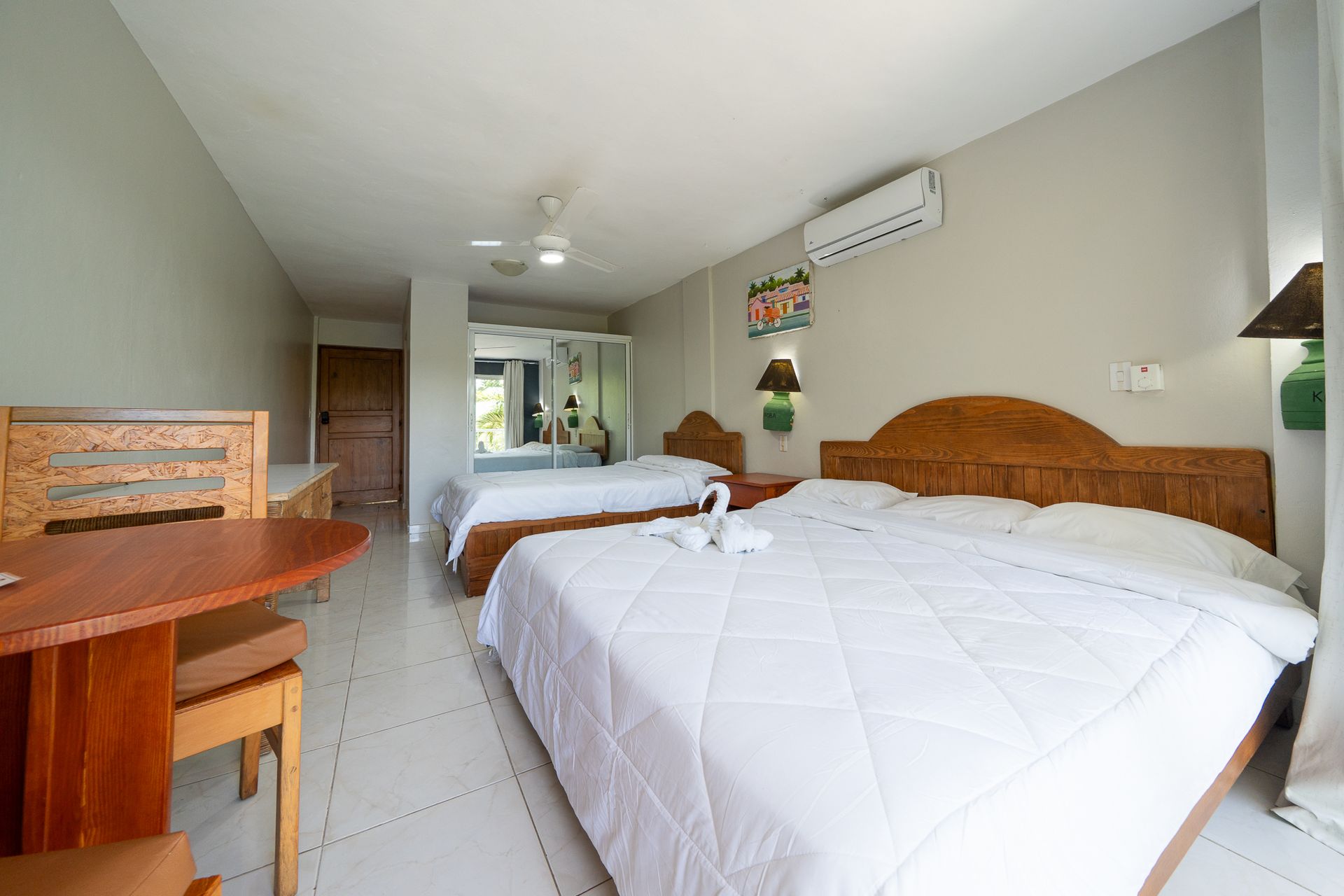 Bright bedroom with two beds, tiled floor, wall-mounted AC, and a small dining table by the window.
