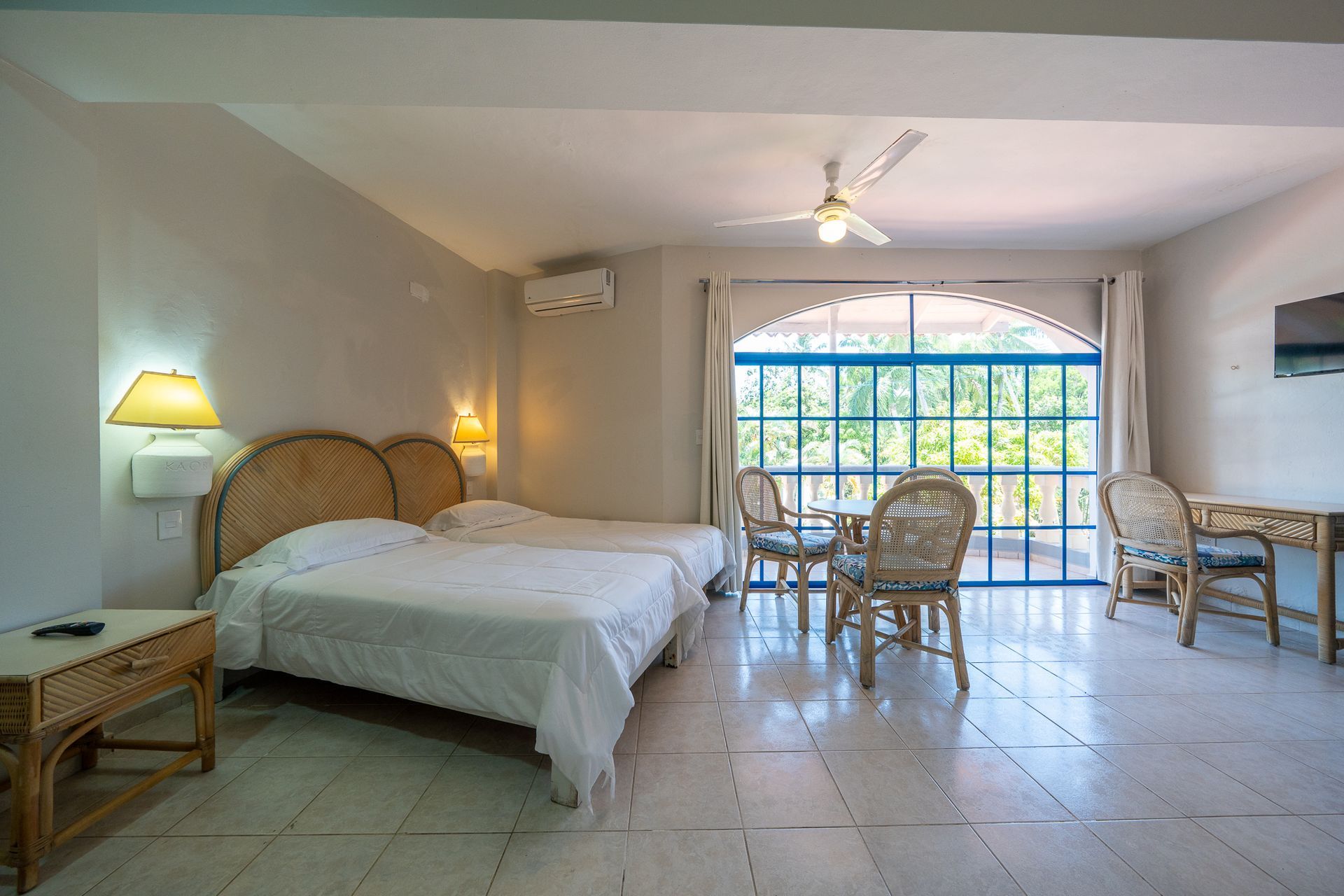 Bright bedroom with twin beds, wicker chairs, tiled floor, and a large window overlooking greenery