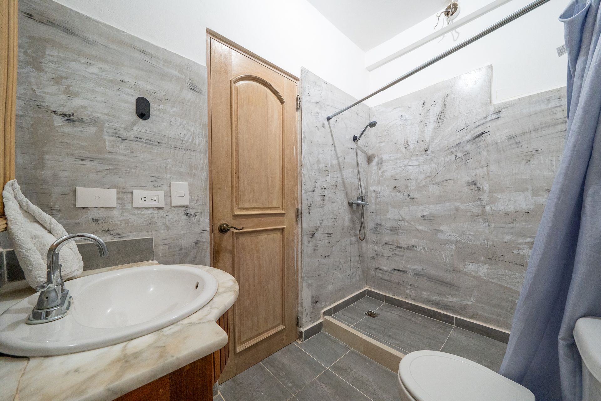 Small tiled bathroom with wooden door, sink vanity, walk-in shower, toilet, and blue towel
