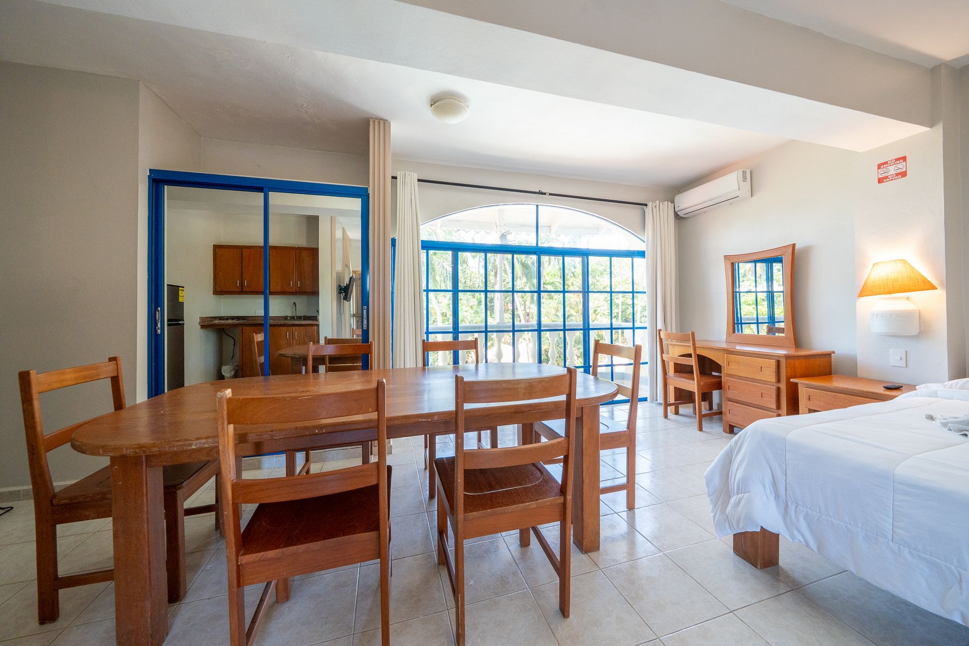 Bright hotel room with wooden tables and chairs, a bed, tiled floor, and large window with blue trim.