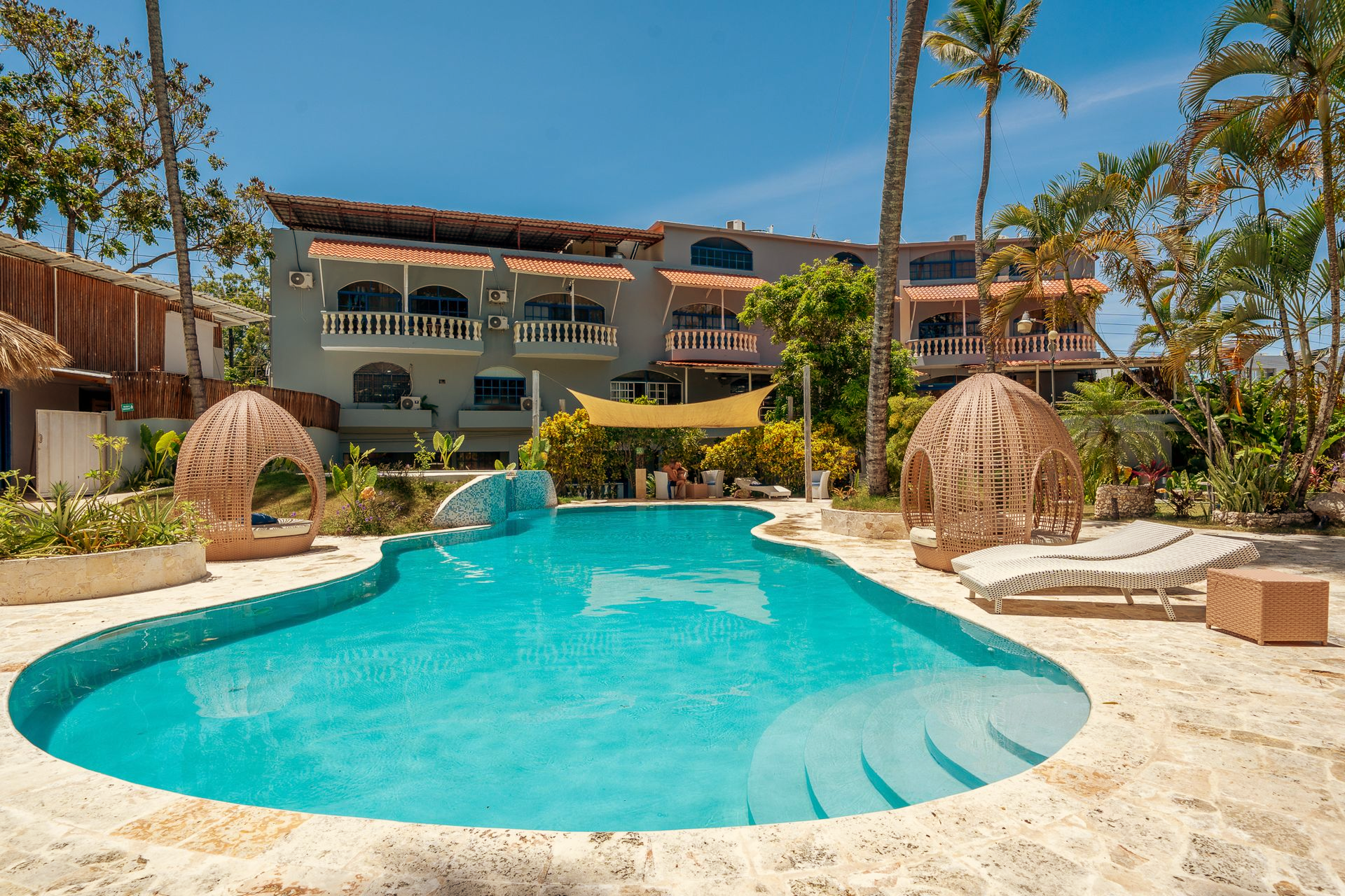 Tropical resort pool with turquoise water, palm trees, and thatched cabana-style loungers near a villa.