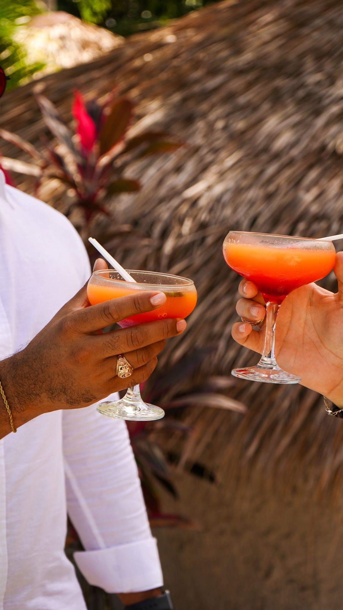 Two people clink orange cocktails in stemmed glasses at a beachside setting.