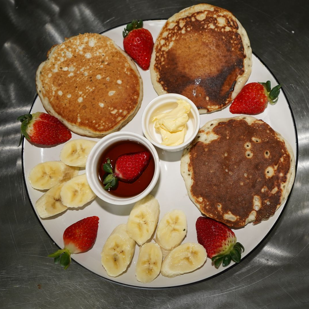 Plate of pancakes with strawberries, banana slices, and small cups of butter and syrup.