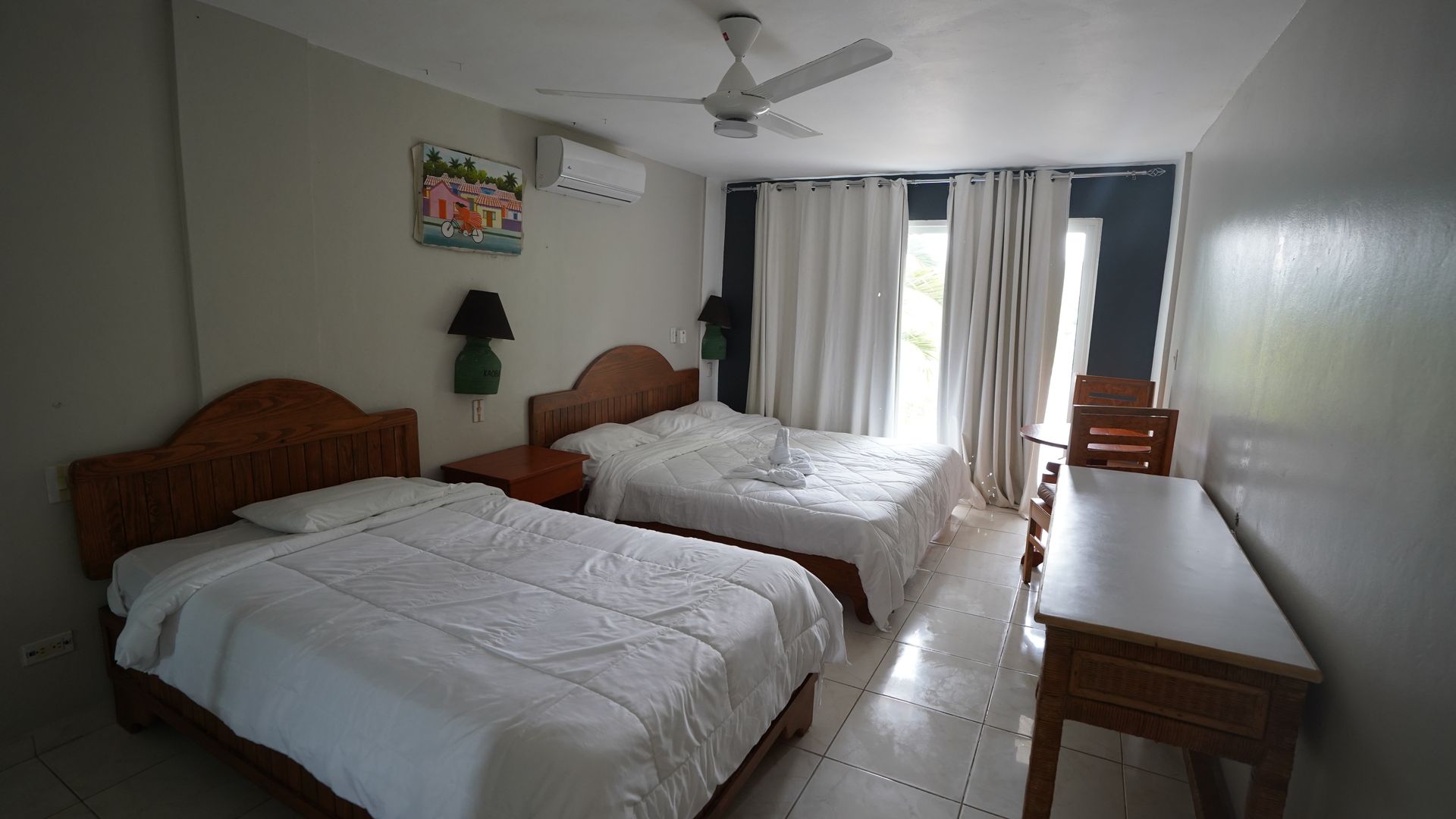 Simple hotel room with two beds, desk, chair, ceiling fan, and curtained window.