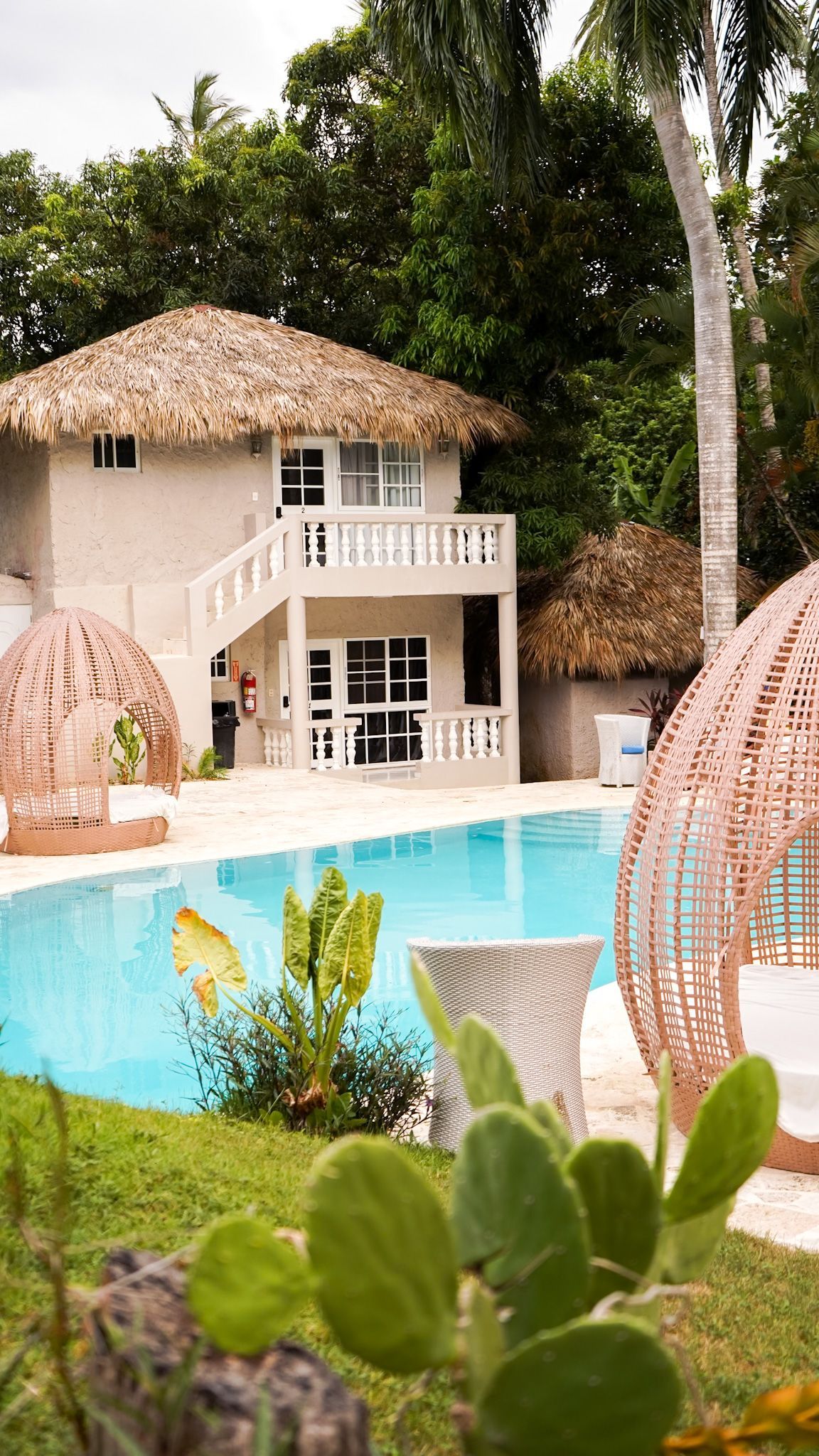 Tropical resort pool with a thatched-roof villa, palm trees, and pink woven chairs beside turquoise water