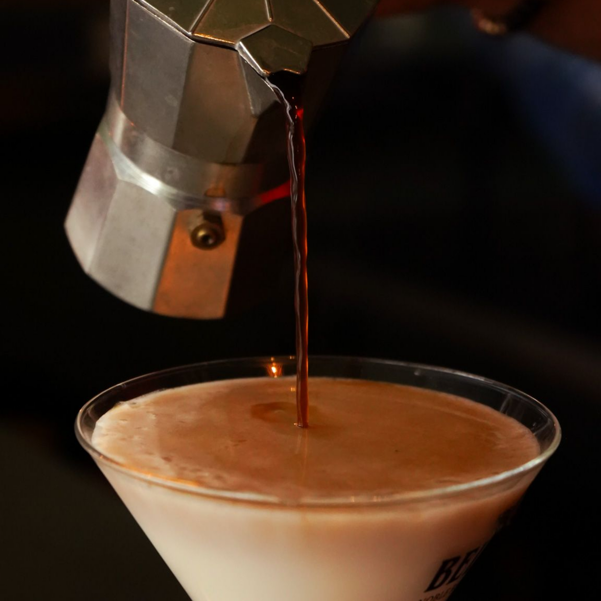 Espresso pouring from a metal spout into a clear cocktail glass with a creamy foam top
