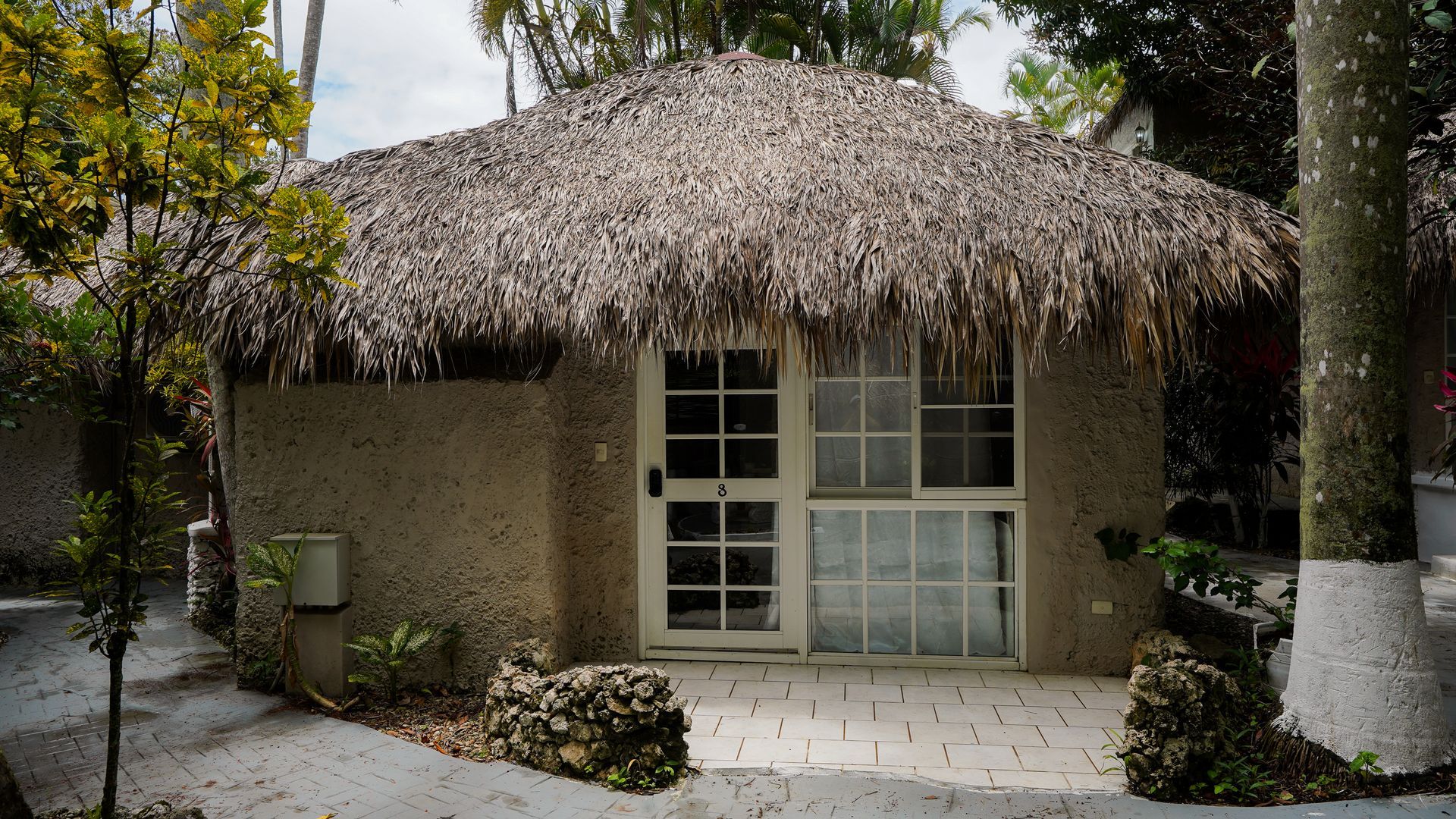 Thatched-roof hut with a white-framed door and windows, set among tropical plants and stone paving