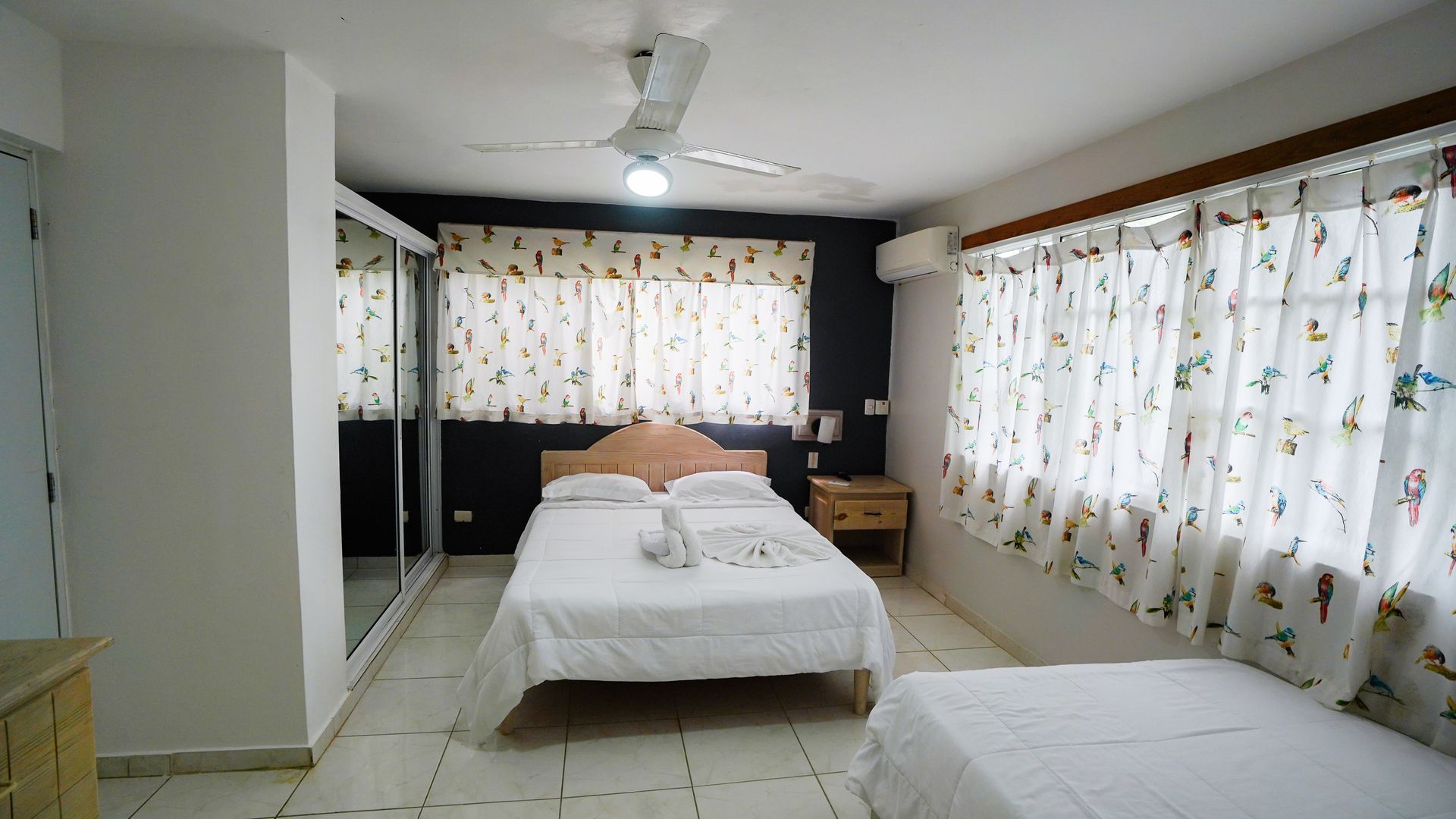 Bright bedroom with two beds, tiled floor, and large windows with sheer floral curtains.