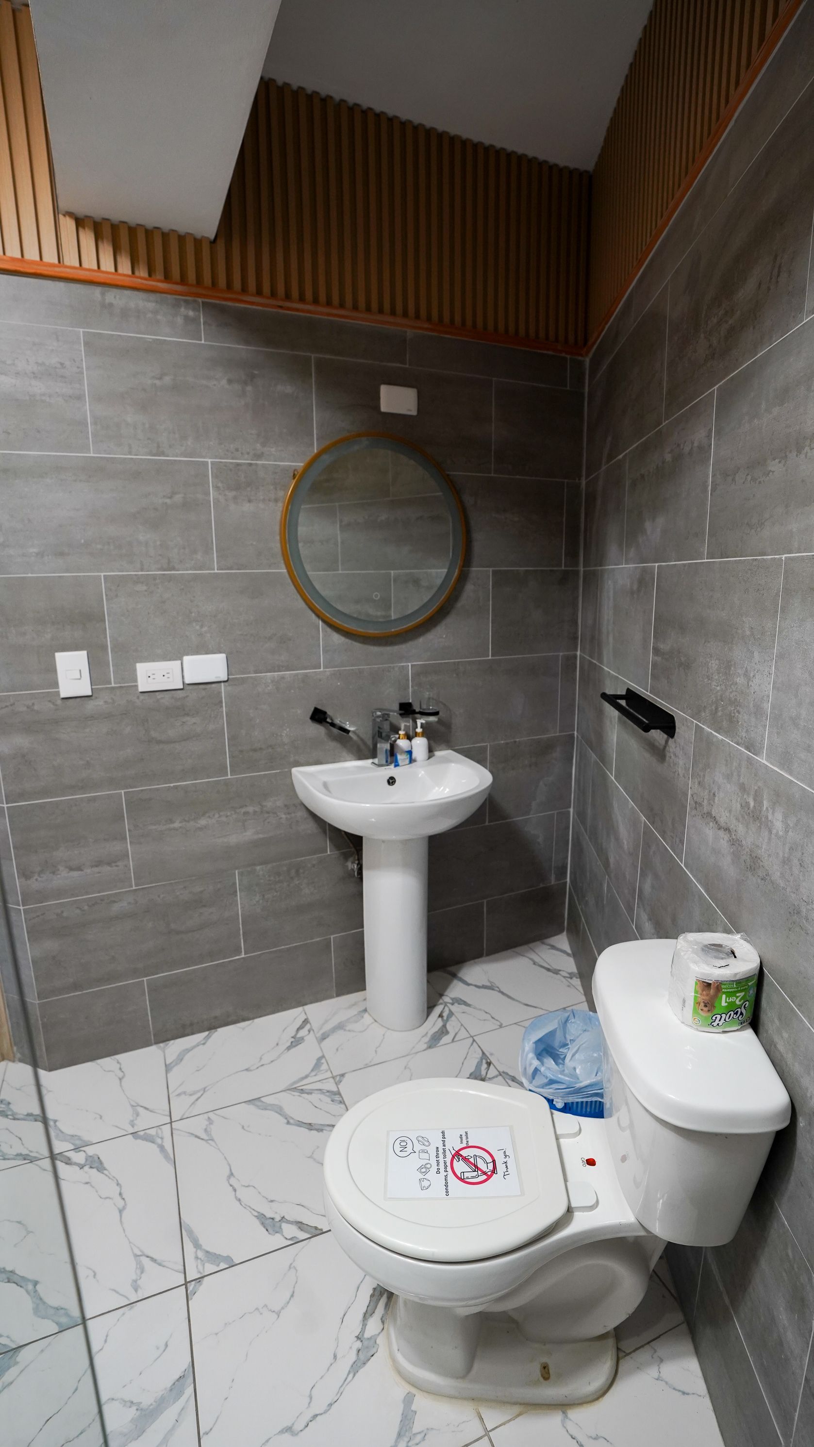 Small tiled bathroom with pedestal sink, round mirror, toilet, and gray walls with wood slats above.