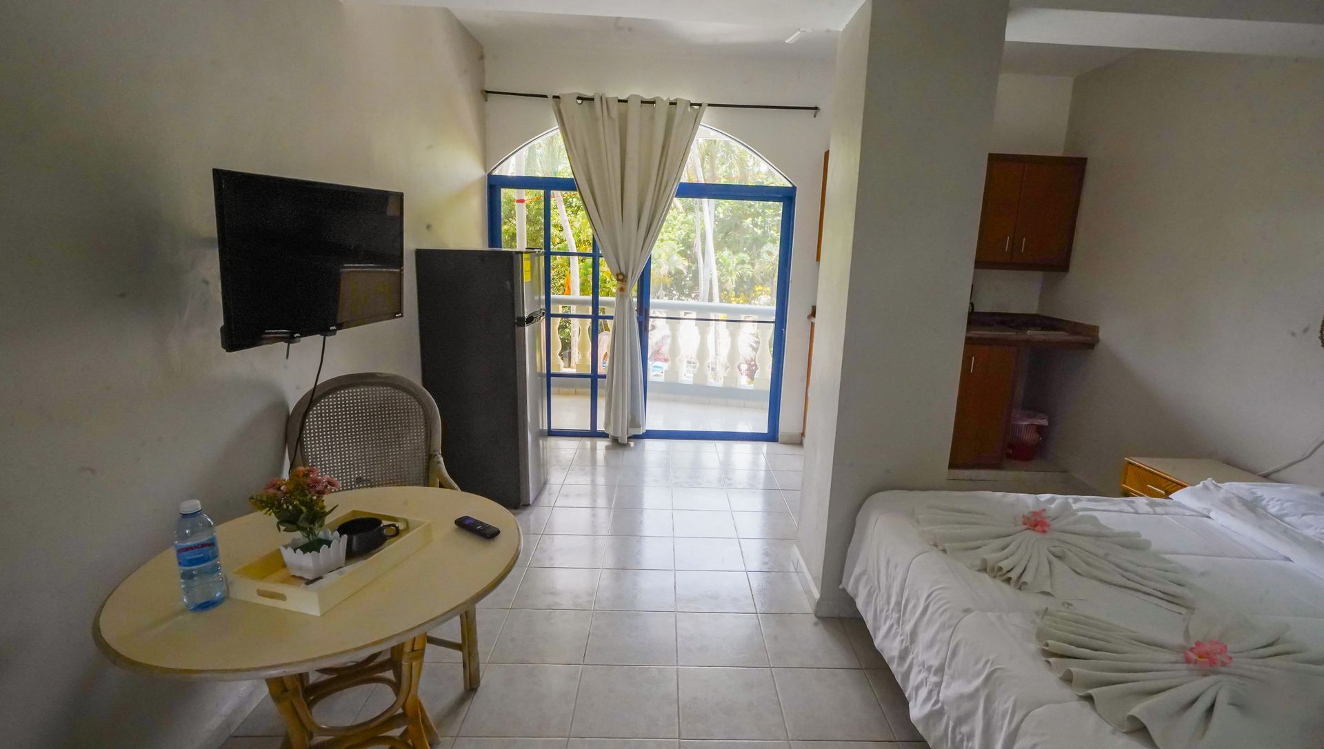 Small studio room with bed, table, TV, chair, and balcony doors letting in daylight