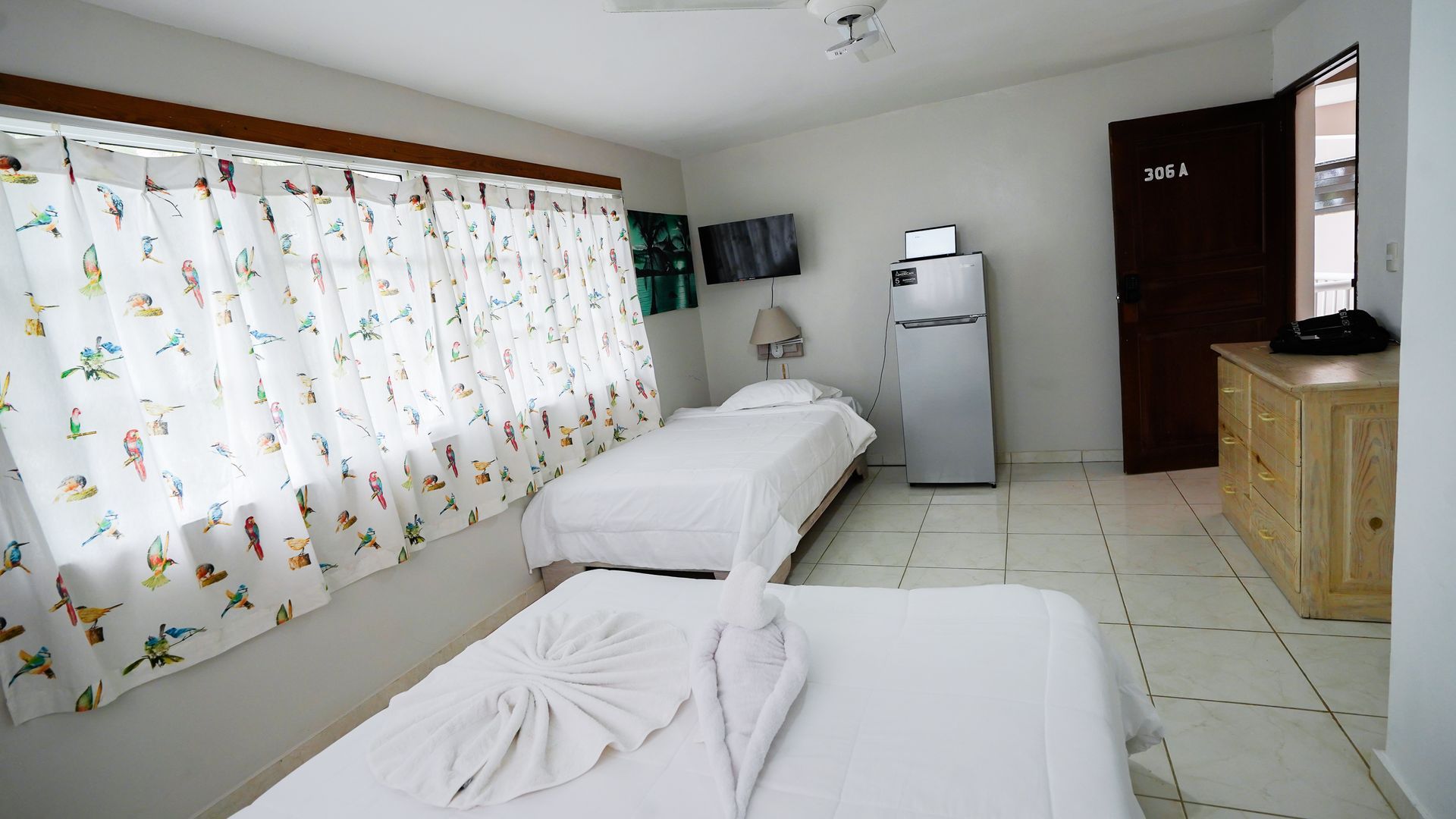 Small bedroom with two beds, a mini fridge, patterned curtains, and a wooden dresser.