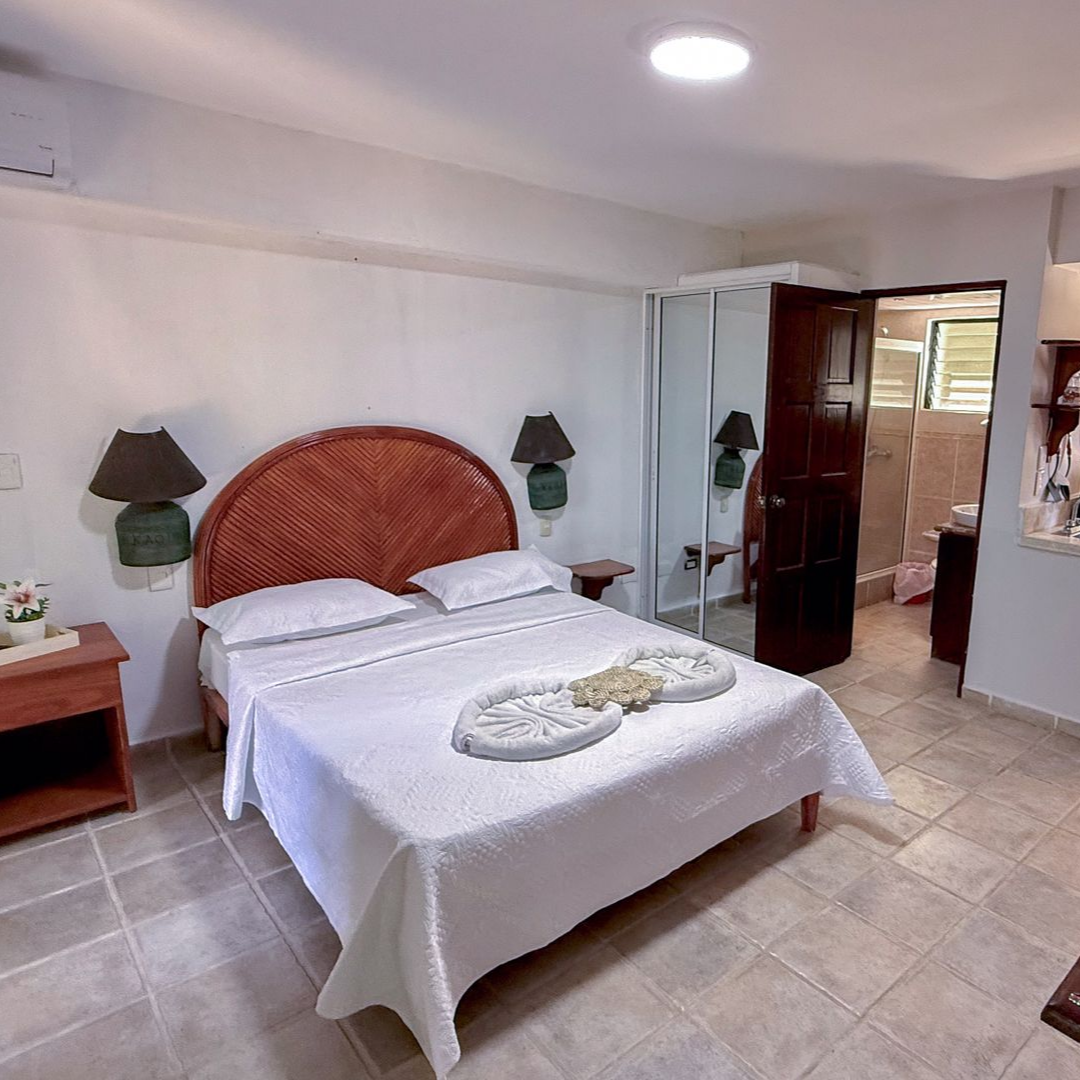 Bright bedroom with a wicker headboard, white bedding, bedside lamps, and an open door to a bathroom.