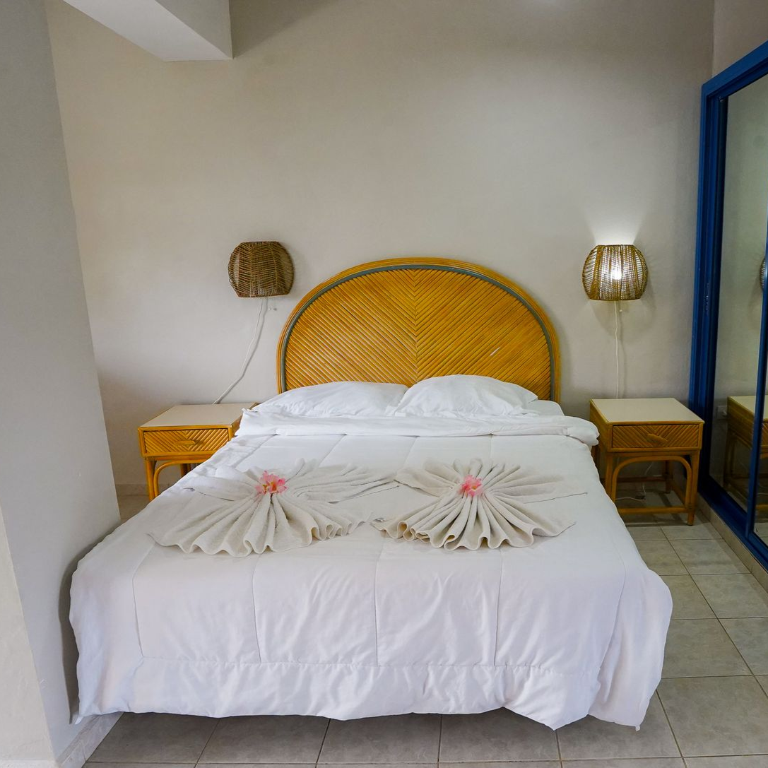 White bed with woven headboard in a simple room, flanked by two wooden nightstands and wall lamps