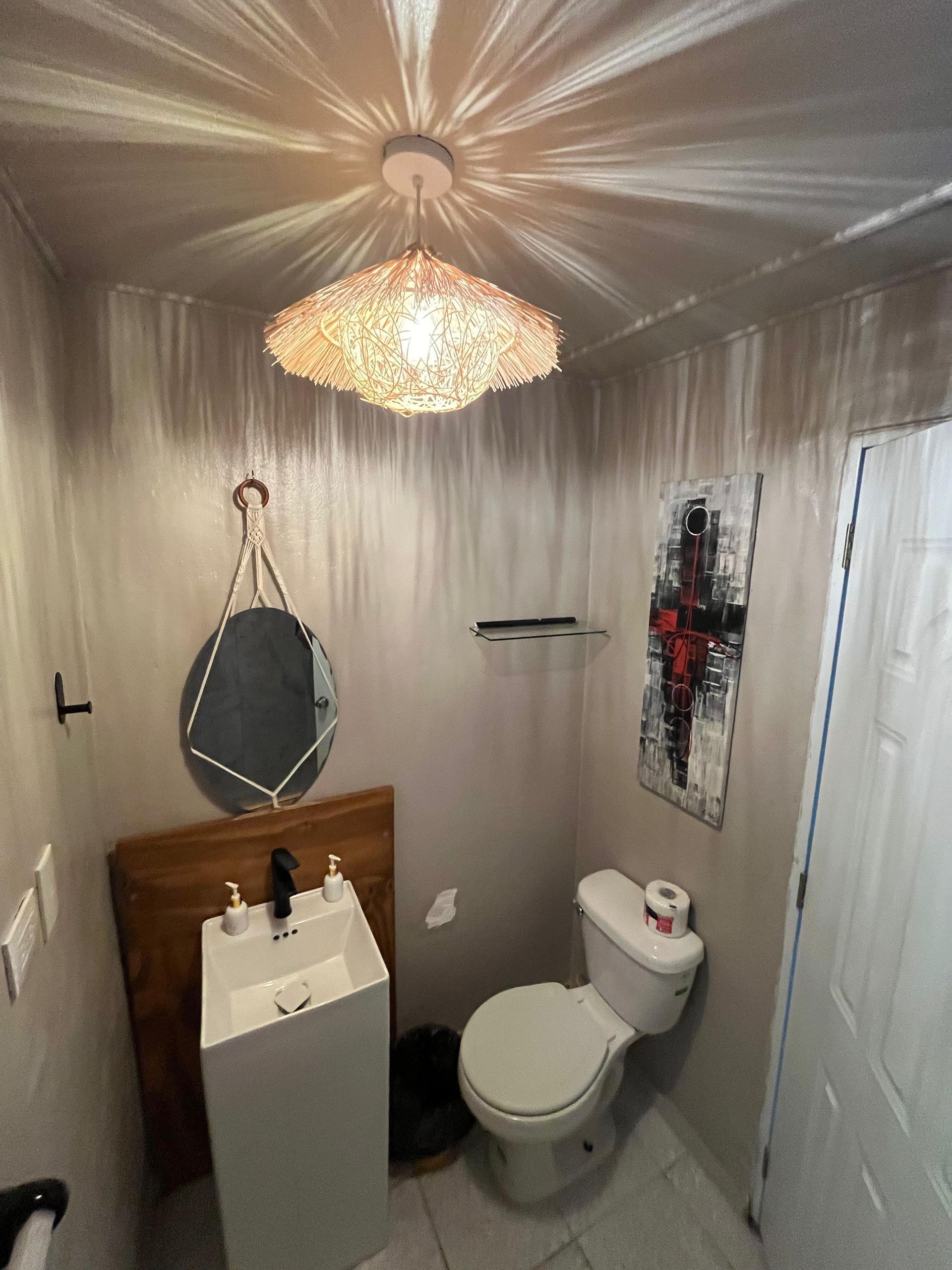 Small bathroom with a toilet, pedestal sink, round mirror, and crystal chandelier under wood-look walls.