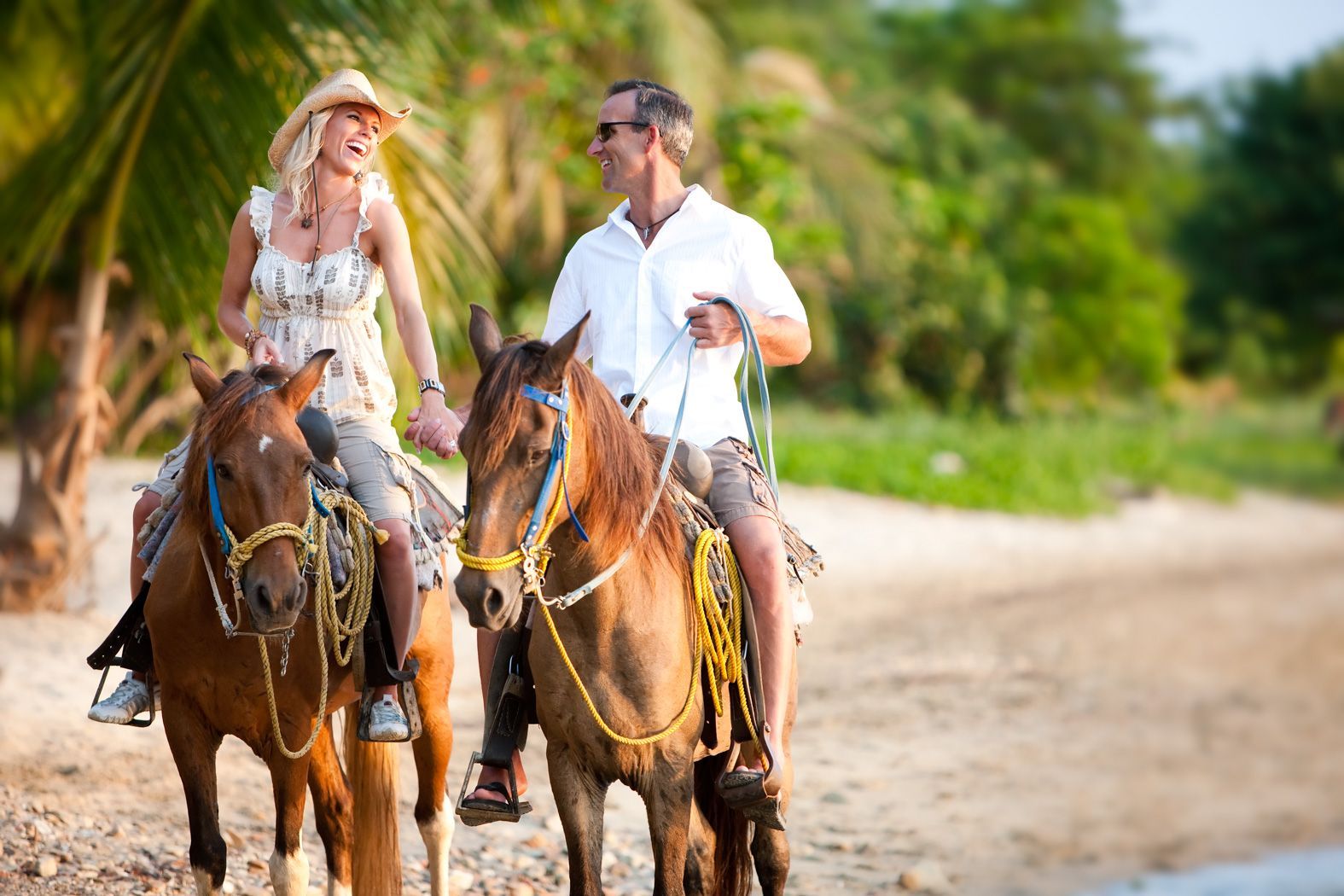 Two people riding horses on a sunny dirt path with palm trees in the background
