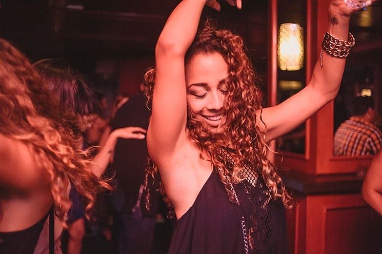 Woman dancing with arms raised in a dimly lit nightclub, smiling under warm red lighting.