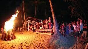 Night beach bonfire with a crowd gathered near a lit wooden platform under palm trees
